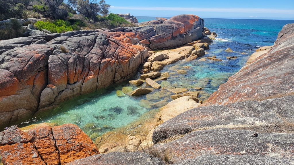 Vibrant orange lichen-covered granite boulders lining the edge of a narrow turquoise water channel at the Bay of Fires, Tasmania.