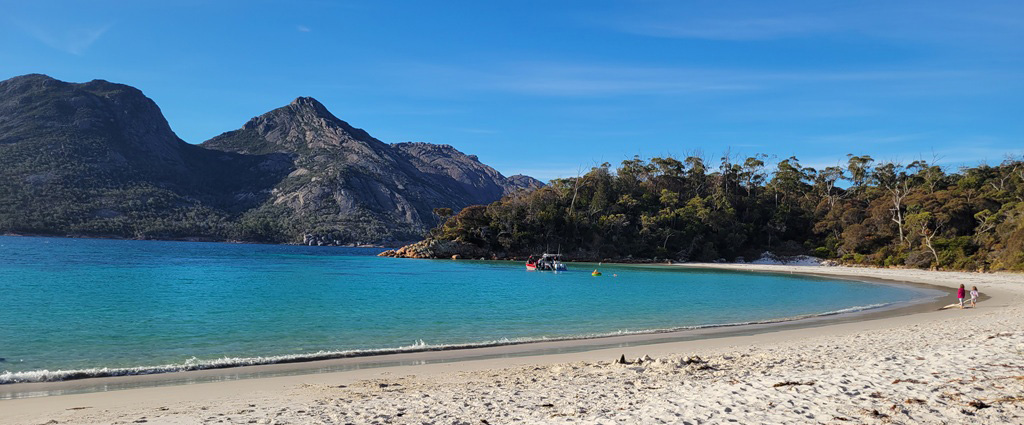 A unique perspective of Wineglass Bay in Freycinet National Park, taken from the white sand beach looking back toward the pink granite peaks of the Hazards under a clear sky.
